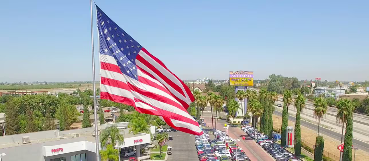 A giant American flag flies over a car dealership with palm trees, a road, and buildings under a clear sky.